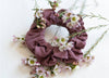 A mauve Kooshoo scrunchie surrounded by flowers on a white background.