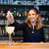 Woman making a cocktail in a bar setting with bottles and decor in the background she is wearing a black gathered headband in her hair
#color_black