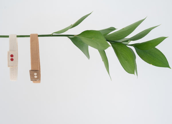 Two Kooshoo hair ties (a brown one and a white one) dangle on a horizontal branch with green leaves.