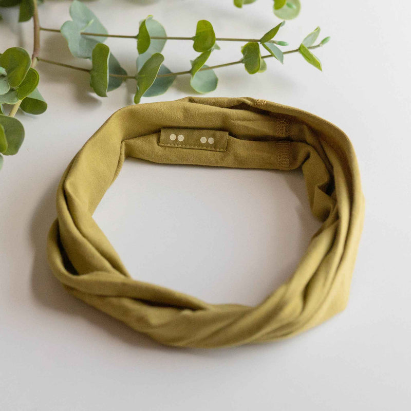 KOOSHOO organic twist headband safari green off-packaging in a flat lay with green leaves #color_willow-green